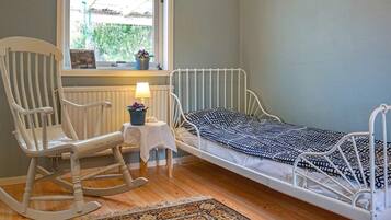 A room with a single bed, a rocking chair, a small table with a lamp, and a window with a view of greenery.