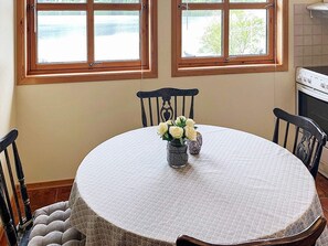 Un comedor con una mesa redonda, dos sillas, un florero con flores blancas y vistas al exterior a través de dos ventanas.