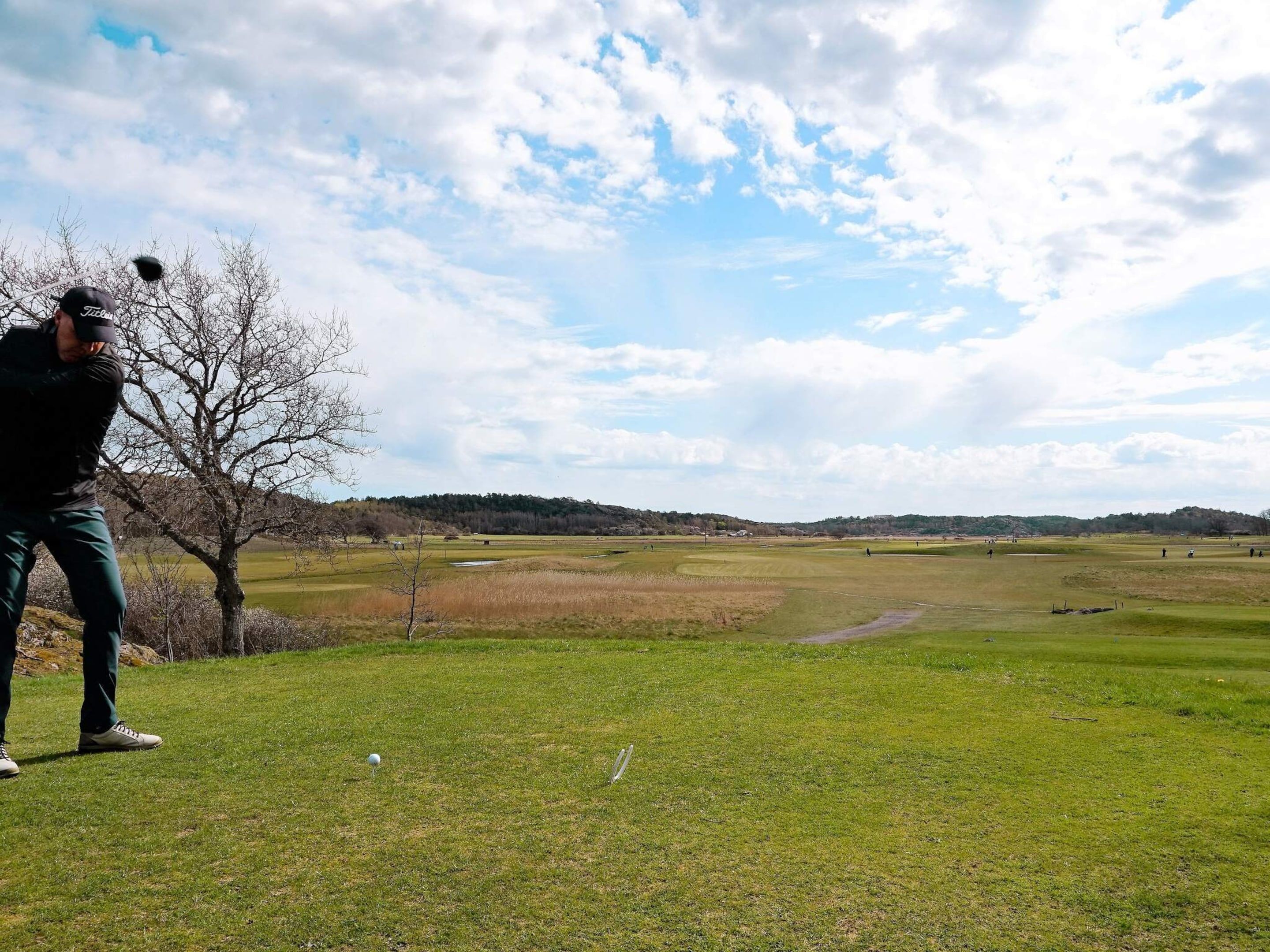 Campo de golfe com céu limpo e um único golfista em primeiro plano.