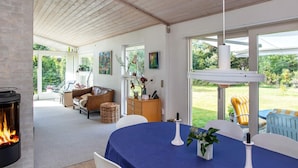 Un salon chaleureux avec une cheminée, une table à manger recouverte d’une nappe bleue et une vue sur le jardin.