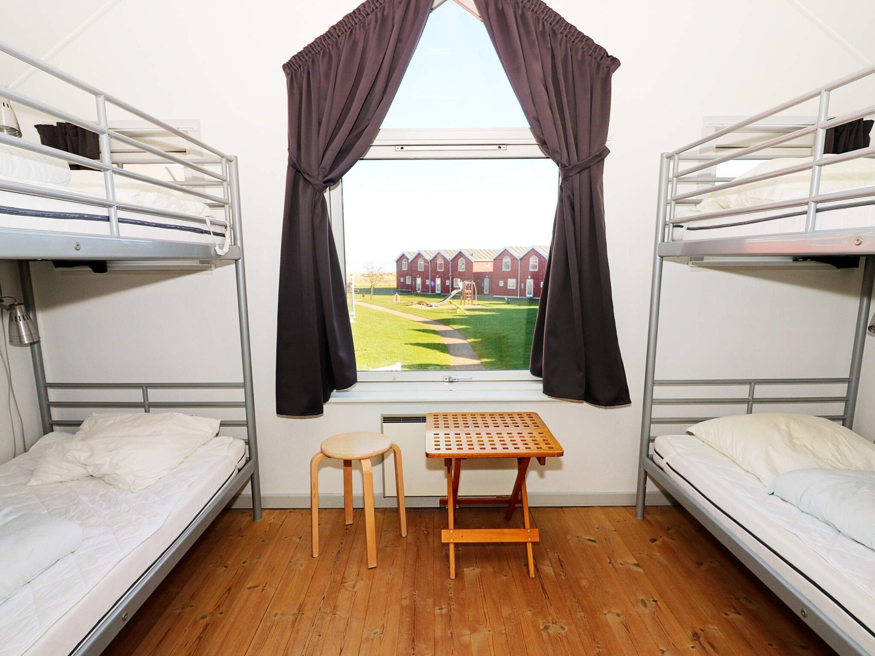 A dormitory room with bunk beds, a small wooden table, and a window with curtains.