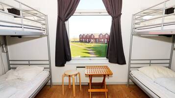 A dormitory room with bunk beds, a small wooden table, and a window with curtains.