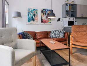 A living room with a grey armchair, a brown leather sofa, and a wooden coffee table. There are two paintings on the wall, a lamp, and a bookshelf with books and decorative items. - 6 Person Holiday Home in Hadsund (Hadsund)