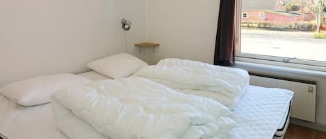 En dobbeltseng med hvide sengetøj i et rum med et vindue, en vægmonteret hylde og en radiator.