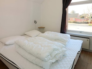 A double bed with white bedding in a room with a window, a wall-mounted shelf, and a radiator.