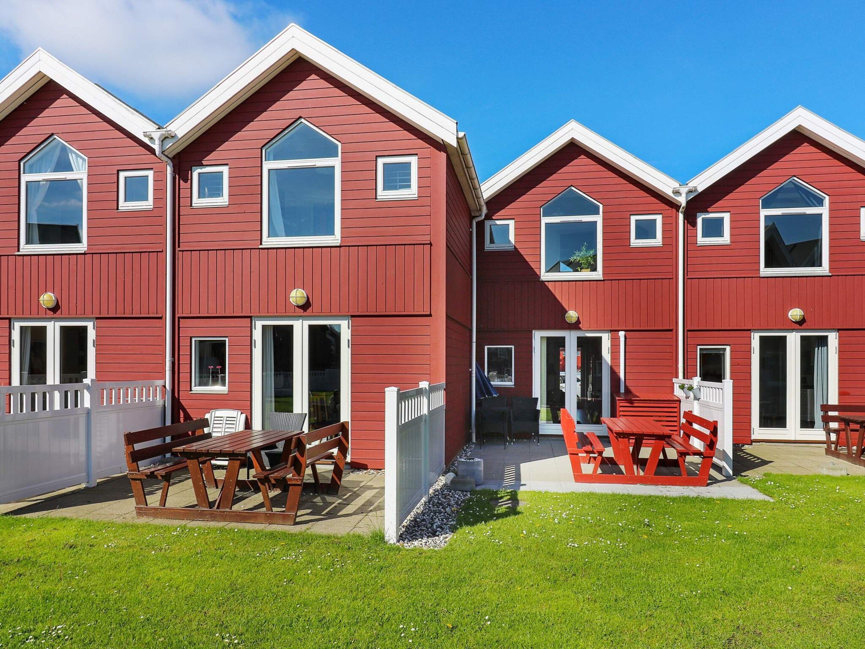 Une rangée de maisons en bois peintes en rouge, avec des boiseries blanches, chacune dotée d’une porte et de fenêtres, ainsi qu’une petite terrasse avec des bancs.