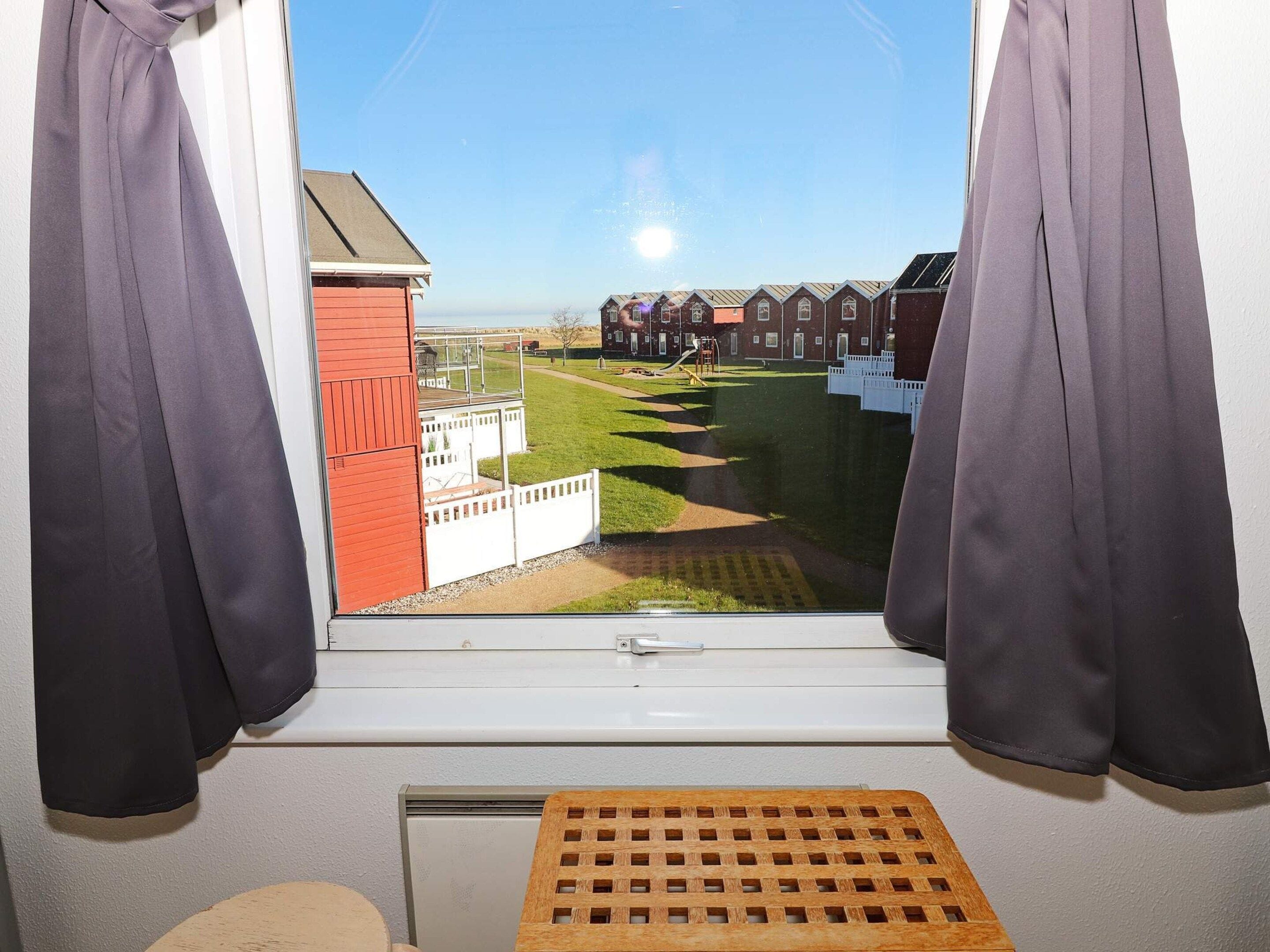Et værelse med vindue der kigger ud over en græsplæne med legeplads, en rød bygning og en række huse.