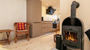 A cozy room with a wooden floor, a wicker chair with a plaid cushion, a modern kitchen with a TV, and a wood-burning stove. - 9 Person Holiday Home in Laeso (Læsø)