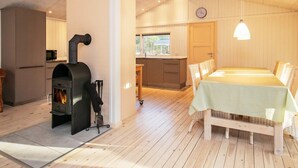 A cozy room with a fireplace, wooden flooring, a dining table with chairs, and a kitchen area with a microwave and a television. - 9 Person Holiday Home in Laeso (Læsø)