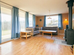 A room with a wooden floor, two sofas, a table, a window with curtains, a wall clock, and a wood-burning stove. - 6 Person Holiday Home in Lokken (Lokken)