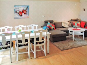 A dining area with a white table and chairs, a grey sofa with colorful cushions, and a small white side table with a glass.