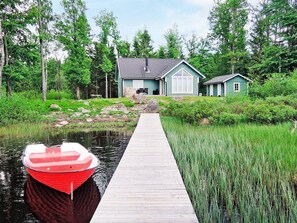 A wooden dock leading to a house with a stone foundation and a green roof, surrounded by lush greenery and a calm body of water.