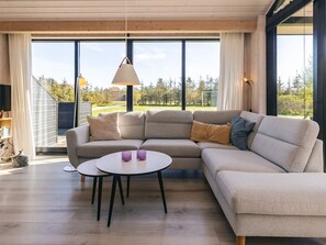 Ein modernes Wohnzimmer mit einer Eckcouch, einem runden Couchtisch und großen Fenstern mit Blick auf eine grüne Landschaft.