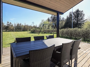 A covered patio with a table and chairs, a swing set, and a view of a grassy field and trees.