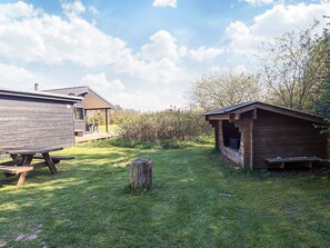 Ein rustikales Blockhaus mit Holzwänden, einem Picknicktisch und einem kleinen Schuppen in einer grünen Umgebung mit Bäumen im Hintergrund.