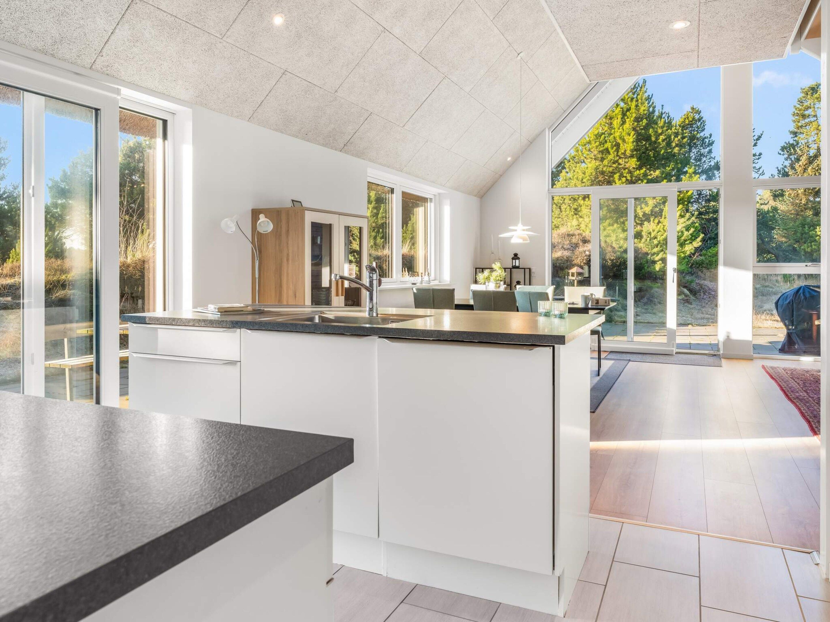 A modern kitchen with a large island, white cabinetry, and large windows offering views of the outdoors.