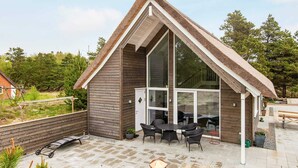 Et moderne hus med stor glasdør, terrasse med udendørs møbler og stråtækt tag.