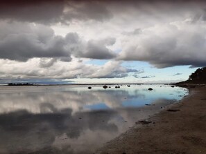 Una playa con agua tranquila que refleja las nubes, un cielo despejado y una costa distante.
