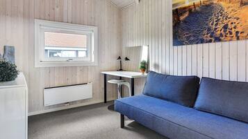 Ein Zimmer mit einer Holzwand, einem Fenster mit Blick auf ein Gebäude, einem Schreibtisch mit Lampe, einem blauen Sofa und einem Bild einer Strandansicht.