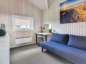 Quarto com parede revestida em madeira, janela com vista para um edifício, escrivaninha com luminária, sofá azul e pintura de uma cena de praia.