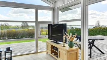 A modern living room with a large TV, wooden furniture, and a view of a green landscape through expansive windows.