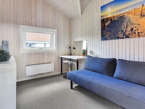 Une pièce avec un mur lambrissé en bois, une fenêtre donnant sur un immeuble, un bureau avec une lampe, un canapé bleu et un tableau représentant une scène de plage.