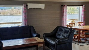 Quarto aconchegante com sofá, mesa de centro e mesa de jantar com cadeiras. Há uma janela com cortinas, um aparelho de ar condicionado na parede e vista para um corpo d'água lá fora.