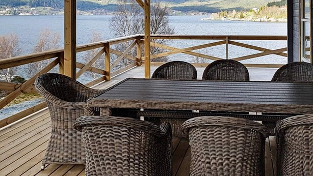 Um terraço coberto ao ar livre com uma mesa e cadeiras de vime, com vista para um lago e montanhas.