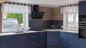 Uma cozinha moderna com armários escuros, fogão embutido e janela com vista para um carro e vegetação lá fora.