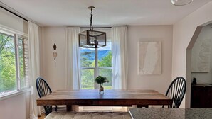 Un comedor con mesa de madera, banco y sillas, una lámpara colgante y vistas al exterior con vegetación.