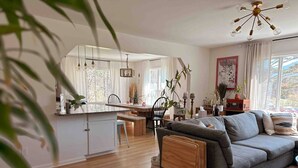 Una sala moderna con un sofá gris, mesitas de madera, un comedor con mesa y sillas, y una cocina con armarios blancos.
