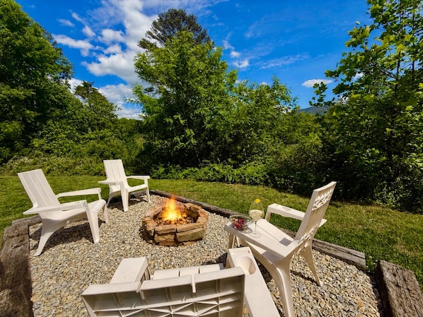 An outdoor seating area with a fire pit, surrounded by white chairs and a table, set on a gravel surface with greenery in the background.