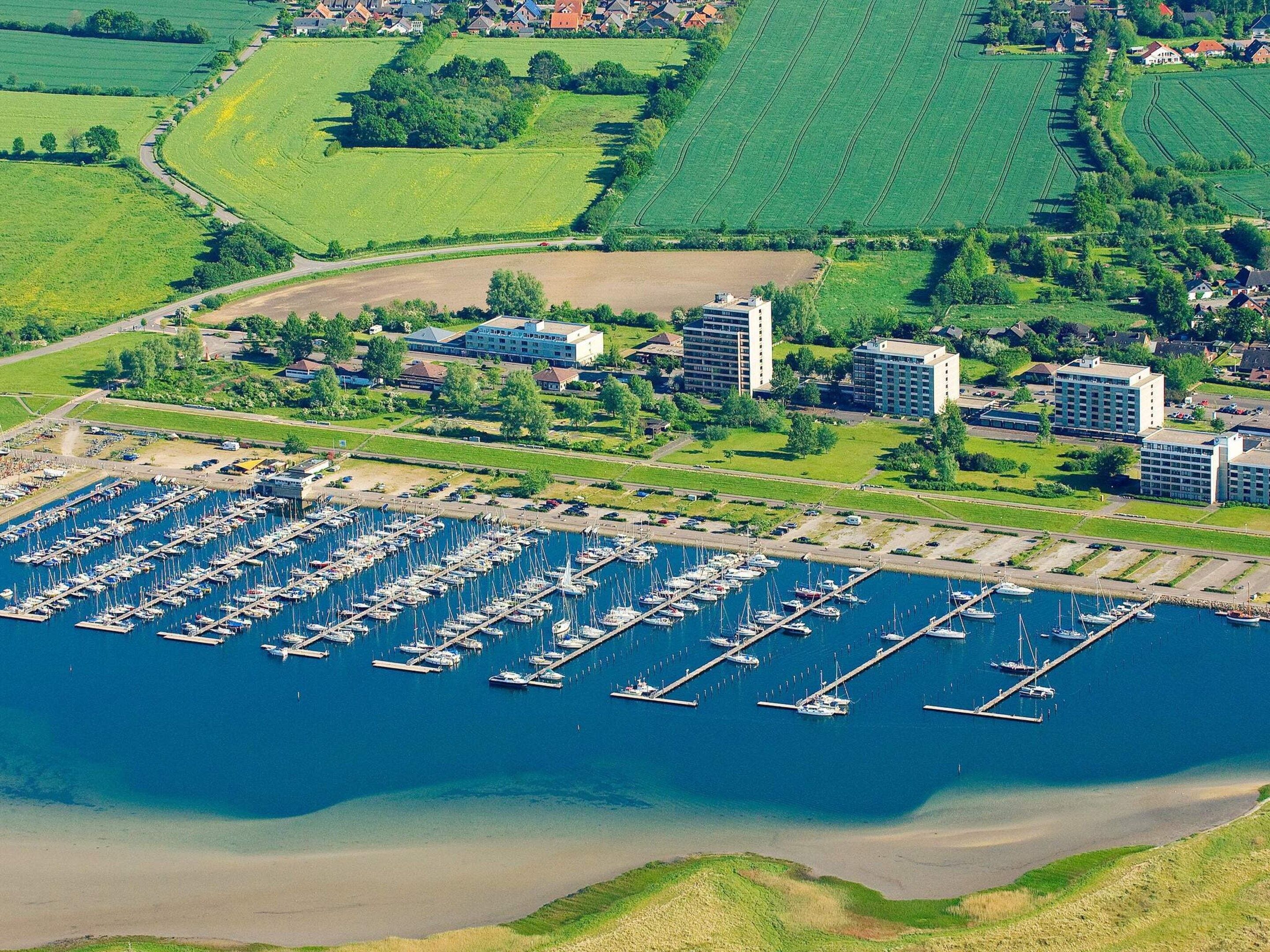 An aerial view of a marina with numerous boats docked, surrounded by green fields and residential buildings.