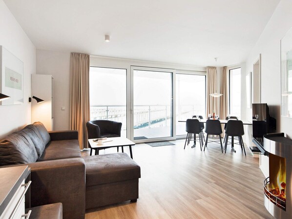 A modern living room with a sofa, a dining table with chairs, a fireplace, and large windows with a view of a bridge. - Exklusive Auszeit mit Meerblick (Wendtorf)