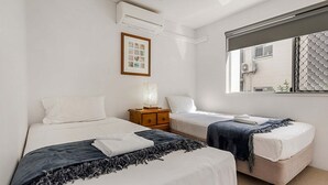 A room with two beds, a small wooden nightstand, a framed picture, and a window with a view of buildings.