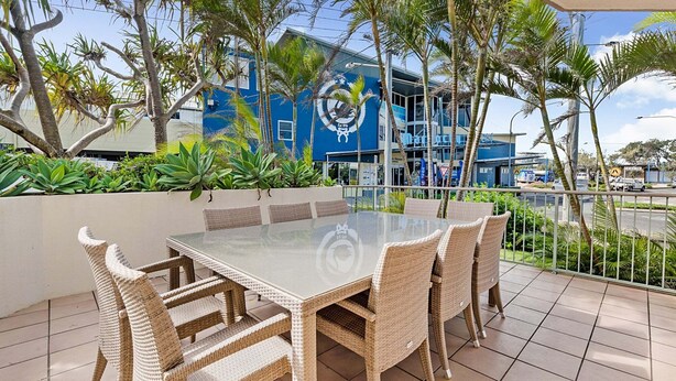 A patio with a glass-top table and wicker chairs, surrounded by tropical plants and overlooking a street with buildings.