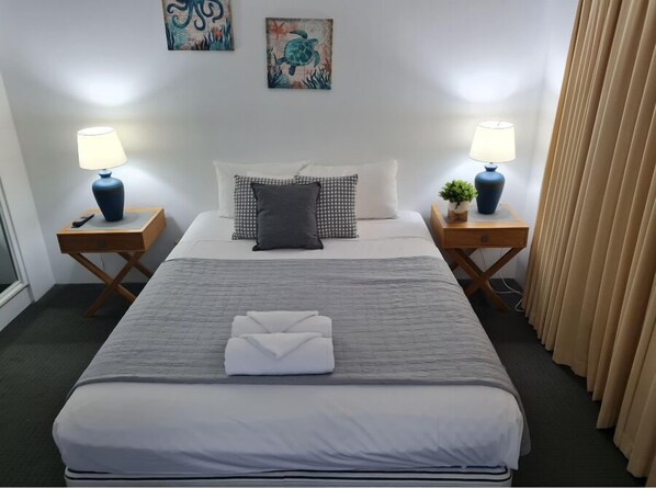 Una cama bien tendida con un edredón gris, dos mesitas de noche con lámparas, una pequeña planta en maceta y una obra de arte en la pared.