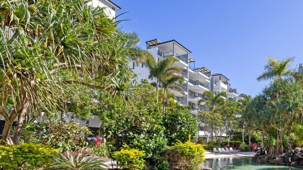 Um resort com piscina cristalina, paisagismo tropical e um edifício com várias andares e sacadas.