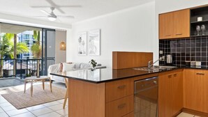 Cozinha moderna com armários de madeira, bancada preta e lava-louças. Há uma área de jantar com sofá e mesa, e uma varanda com vista para palmeiras.