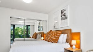 Quarto com uma cama grande, mesa de cabeceira com abajur, uma cadeira e porta de vidro corrediça que dá para uma varanda.