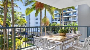 Varanda com mesa de madeira e cadeiras brancas, vista para um jardim com palmeiras e um edifício ao fundo.