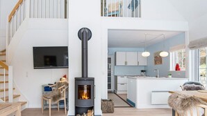 En moderne stue med en centralt placeret brændeovn, et fjernsyn monteret på væggen og en trappe, der fører til en etage ovenpå.