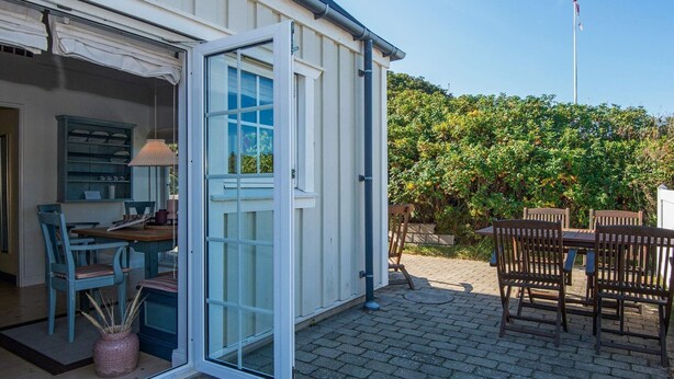 Et hus med terrasse, hvor der er et bord og stole, en dør der fører ind, og en have med en hæk.