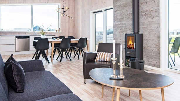 A modern living room with a fireplace, a dining area with a wooden table and chairs, and a kitchen with a dishwasher and large windows.