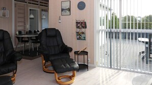 Een kamer met twee zwarte relaxfauteuils, een kleine tafel, een eethoek met stoelen, een houten vloer, een groot raam met jaloezieën en een wandklok.