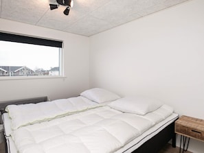 A single bed with a white mattress and pillows, a wooden bedside table, a window with a view of buildings, and a ceiling light fixture. - 5 Star Holiday Home in Bogense (Bogense)