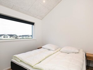 A single bed with a white mattress and pillows, a wooden headboard, and a window with a view of houses.