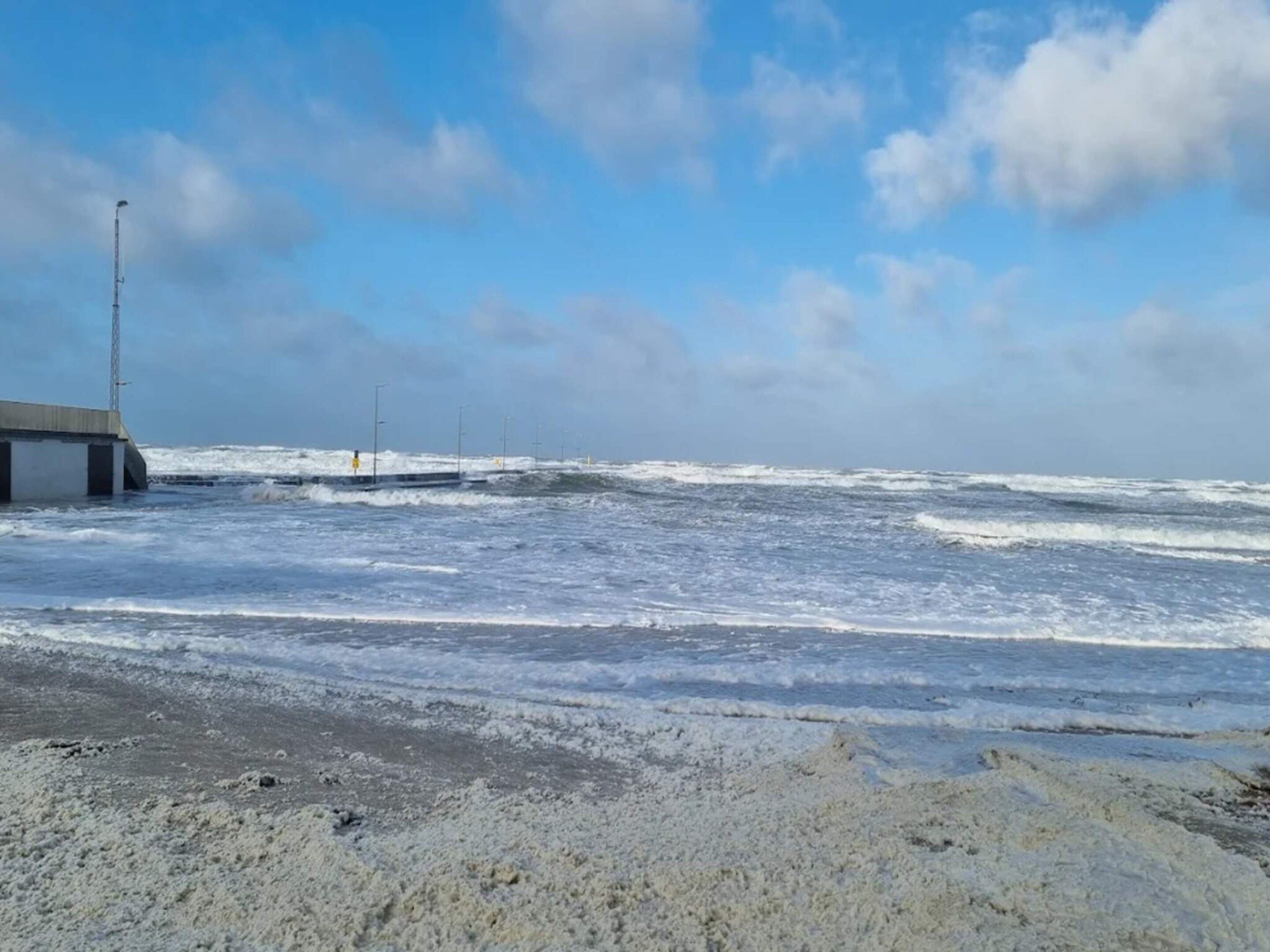 Una playa con olas rompiendo en la orilla, un muelle que se adentra en el agua y un cielo despejado.