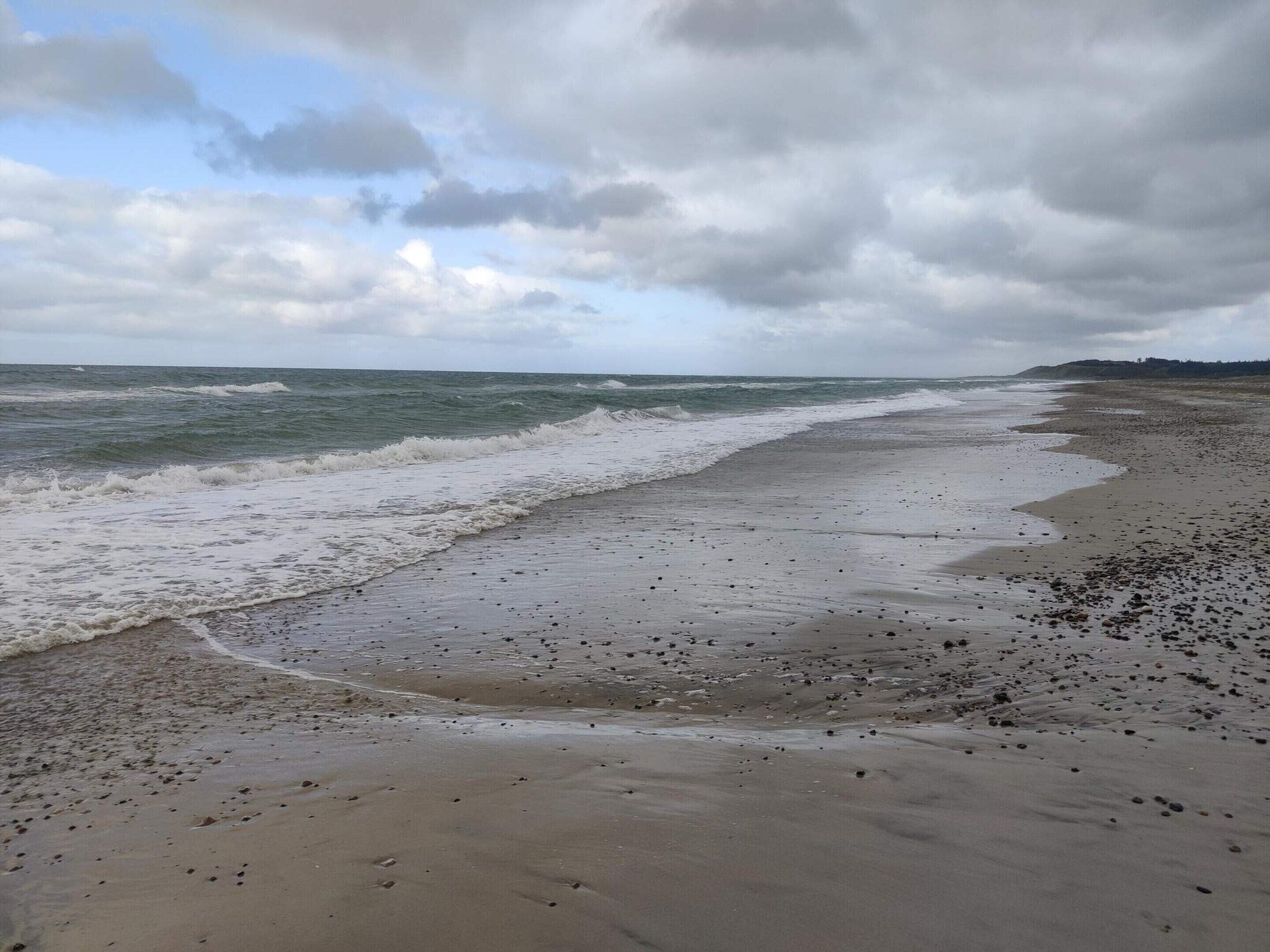 A beach with waves, pebbles, and a cloudy sky.