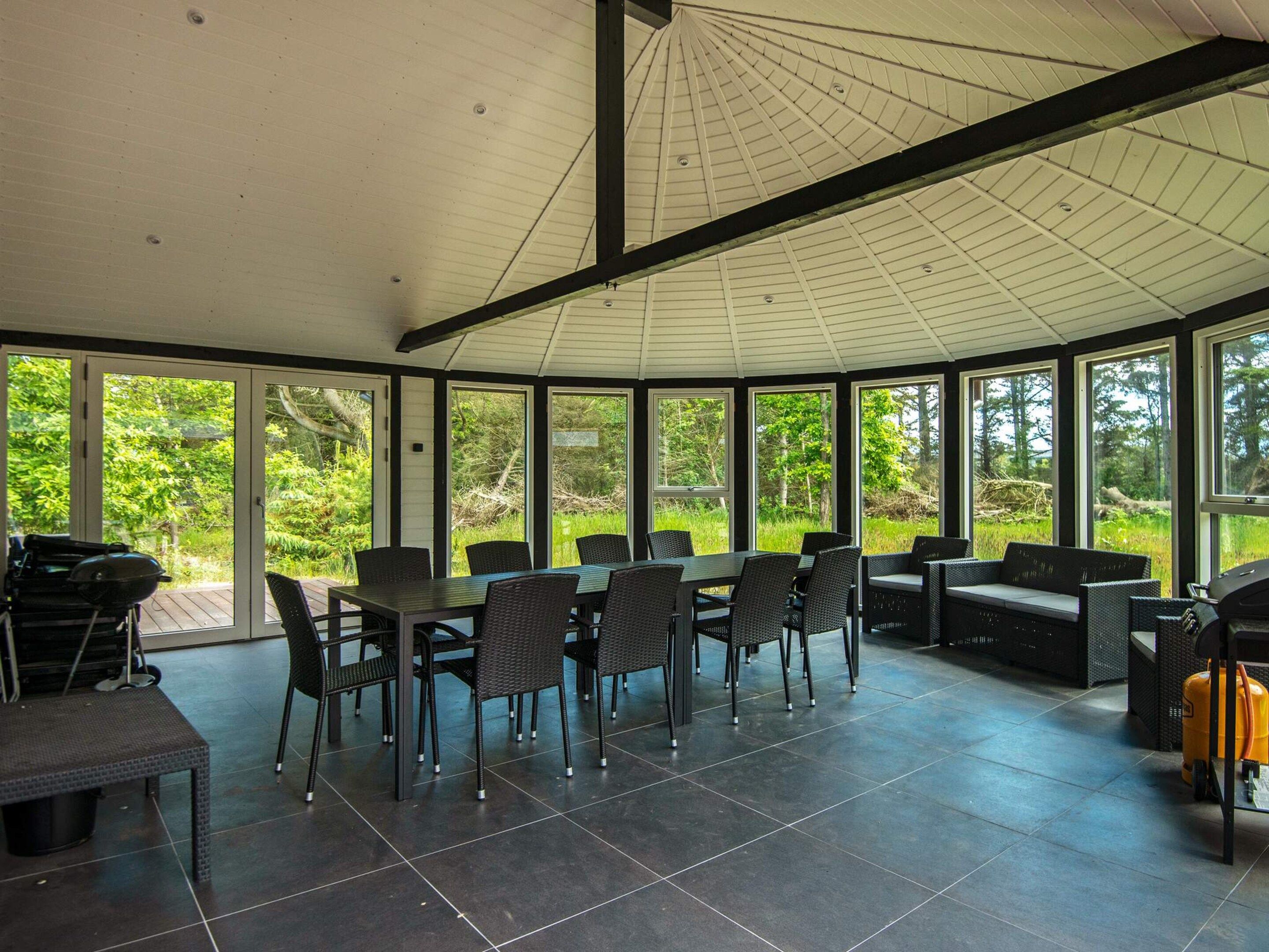 A covered outdoor dining area with a table and chairs, a grill, and a view of the surrounding greenery.